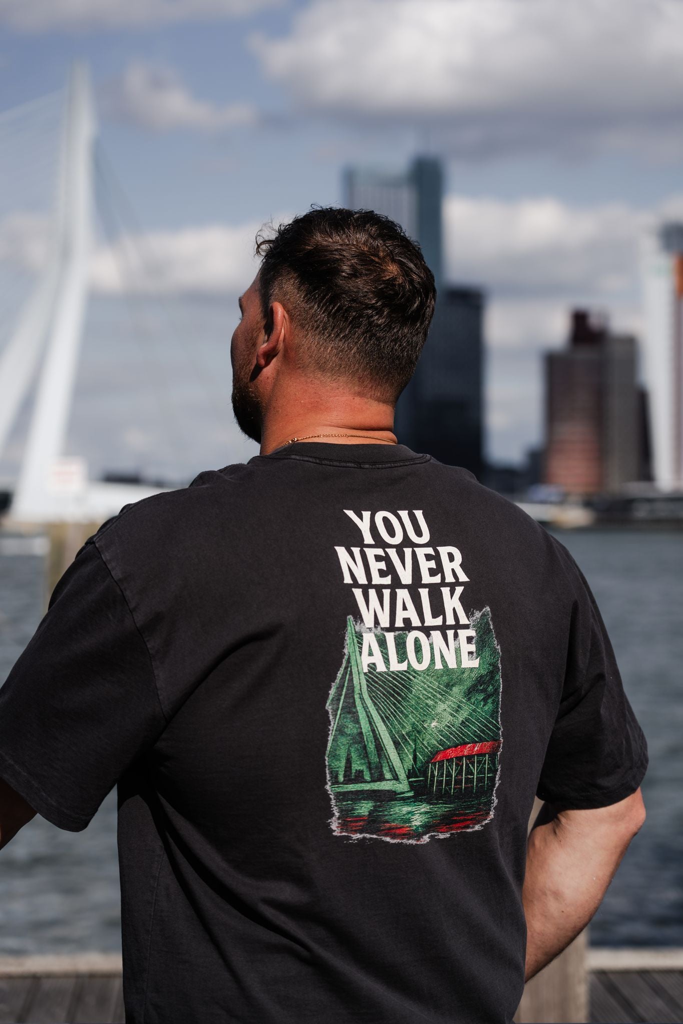 Oversized T-shirt met opdruk “YOU NEVER WALK ALONE” en illustratie van De Kuip, Erasmusbrug en skyline van Rotterdam in groen-rood kleurenpalet.
