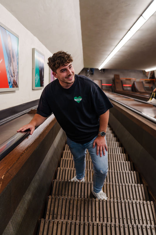 Groen-wit hartvormig TRØBL-logo als borstborduring op zwart oversized T-shirt, geïnspireerd op de stad Rotterdam.